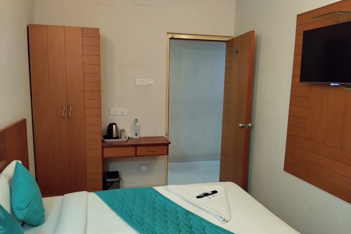 Habitación de hotel con cama, armario, escritorio con hervidor, cesto de basura y televisor montado en la pared.