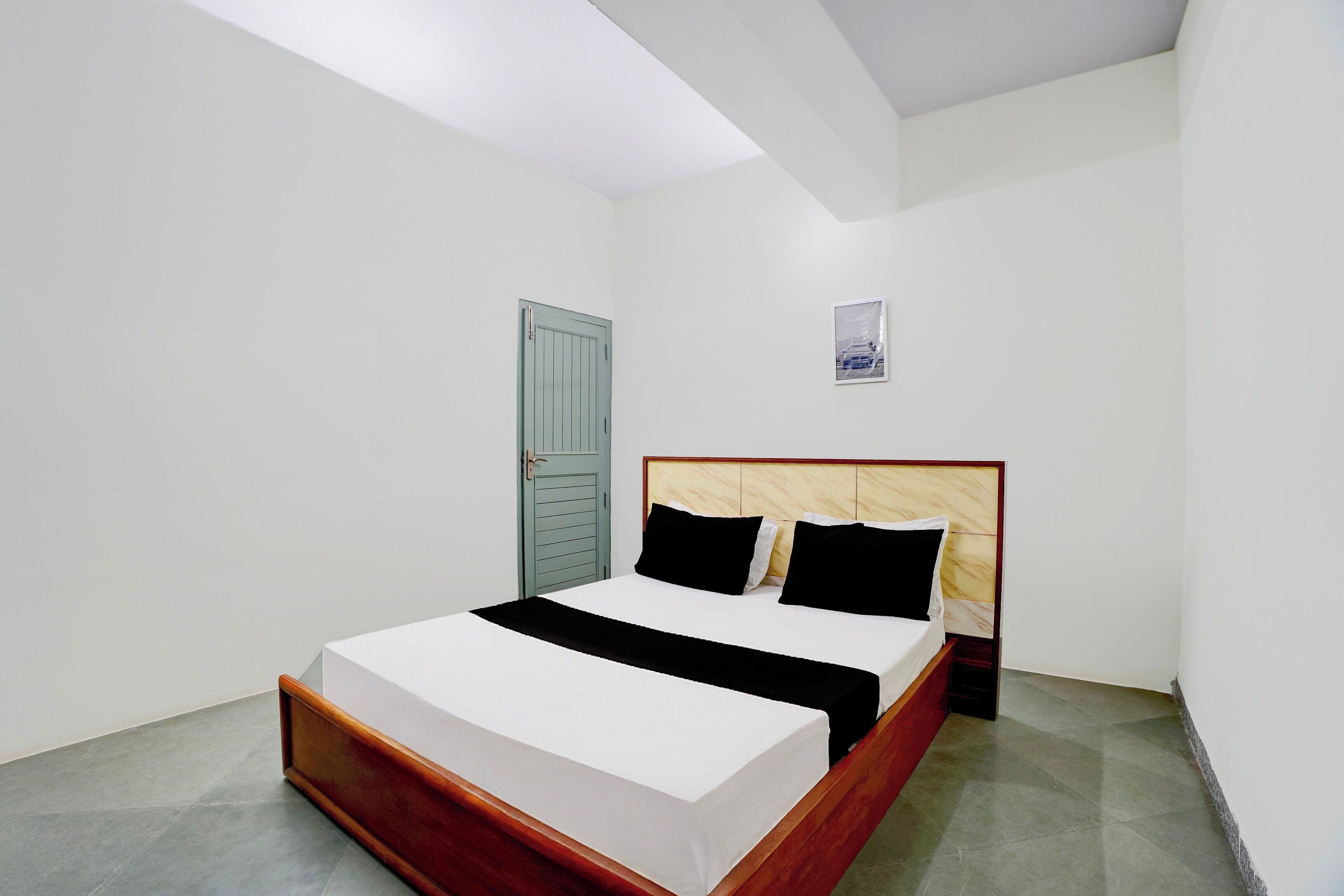 Una cama individual con estructura de madera y ropa de cama blanca en una habitación con paredes blancas y suelo gris.