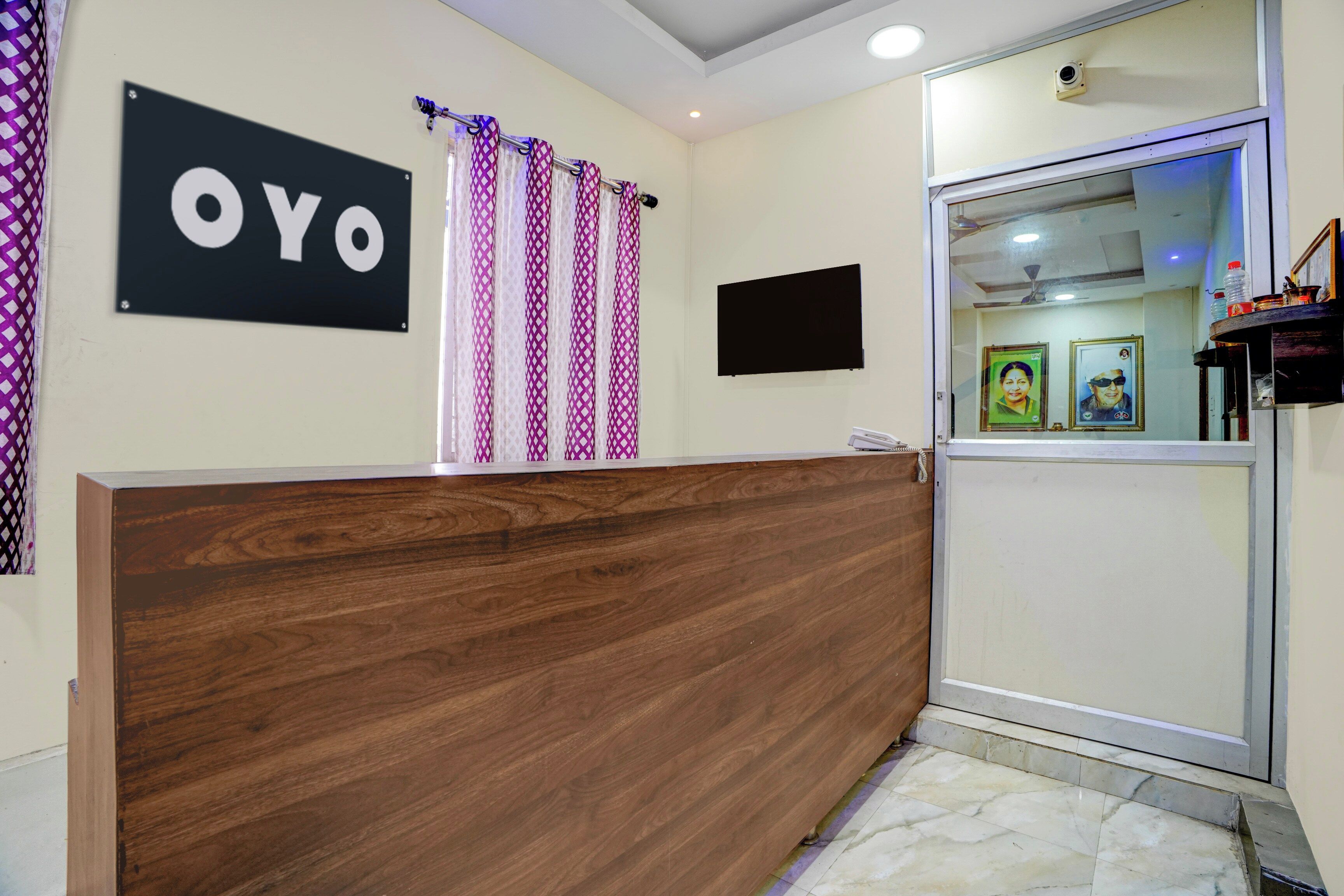 A reception desk with a wooden finish, a sign with the word 'OYO', patterned curtains, a flat-screen TV, and framed pictures on the wall.