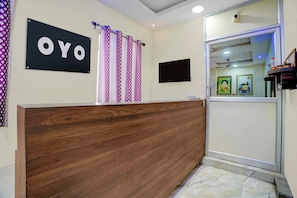 Un mostrador de recepción con detalles de madera, un cartel con la palabra 'OYO', cortinas con estampado, un televisor de pantalla plana y cuadros enmarcados en la pared.