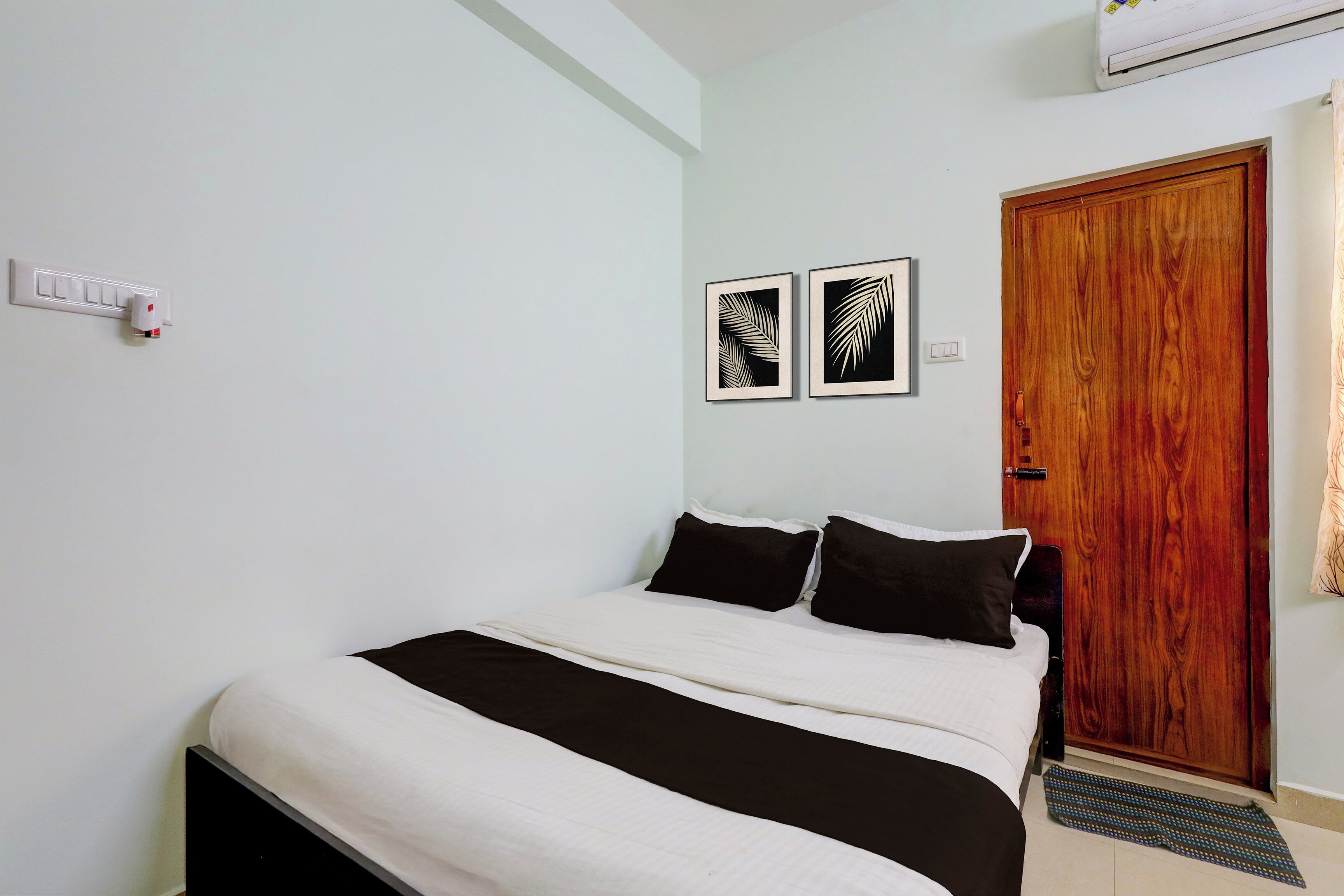 A bedroom with a bed, two wall-mounted light switches, a wooden door, and two framed wall art pieces.