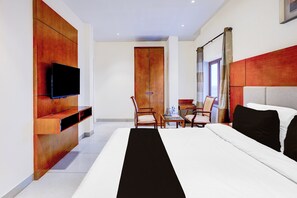 A hotel room with a bed, a TV mounted on the wall, a wooden wardrobe, a small table with chairs, and a window with curtains.