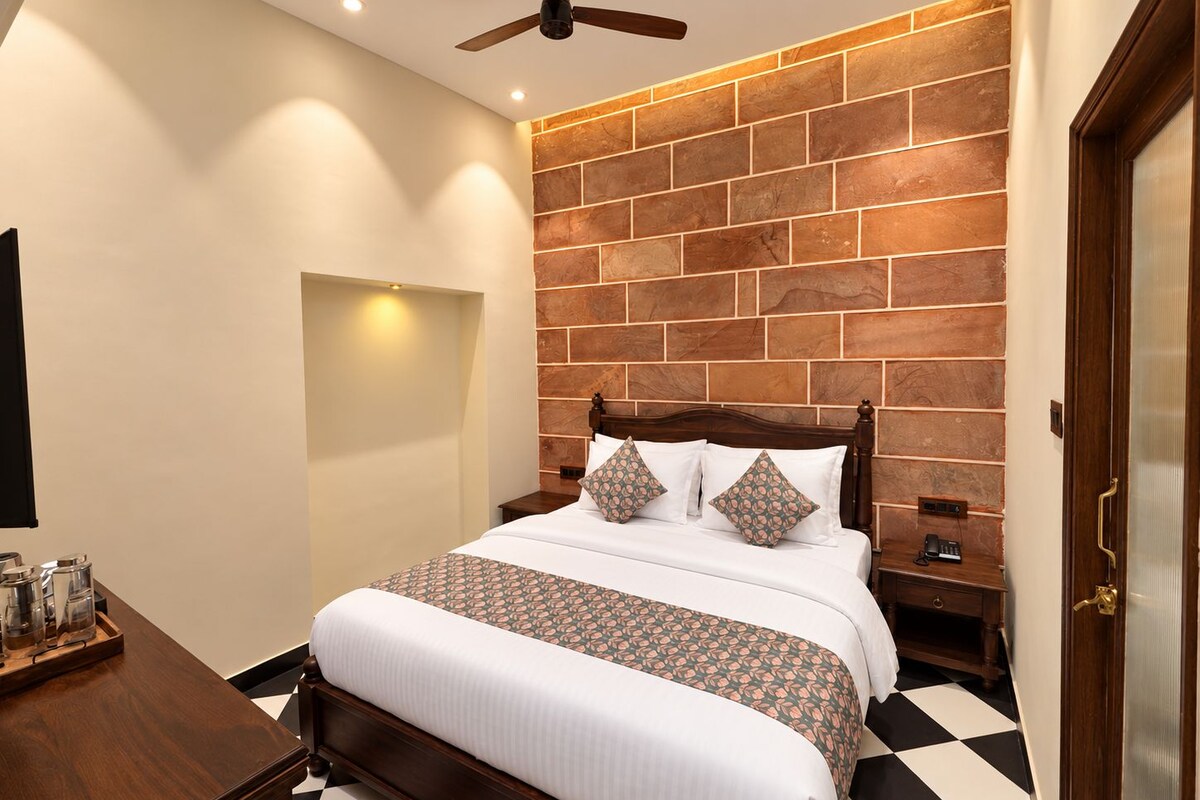 Une chambre d’hôtel avec un mur au motif de briques, un lit en bois avec une literie blanche, un ventilateur de plafond et un sol à carreaux noirs et blancs.