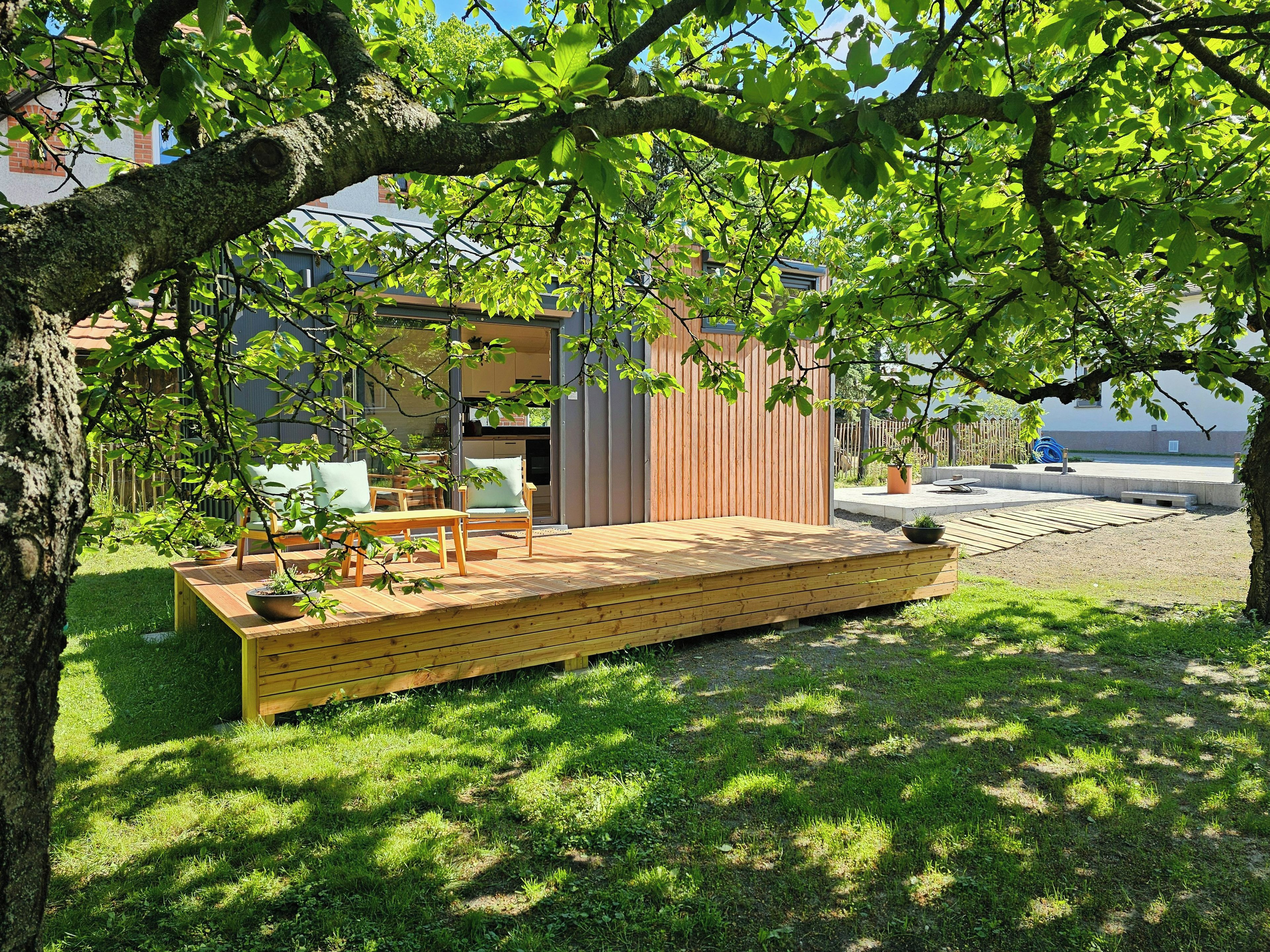 Eine Holzterrasse mit Gartenmöbeln, ein kleiner Gartenbereich und ein Gebäude im modernen Stil.