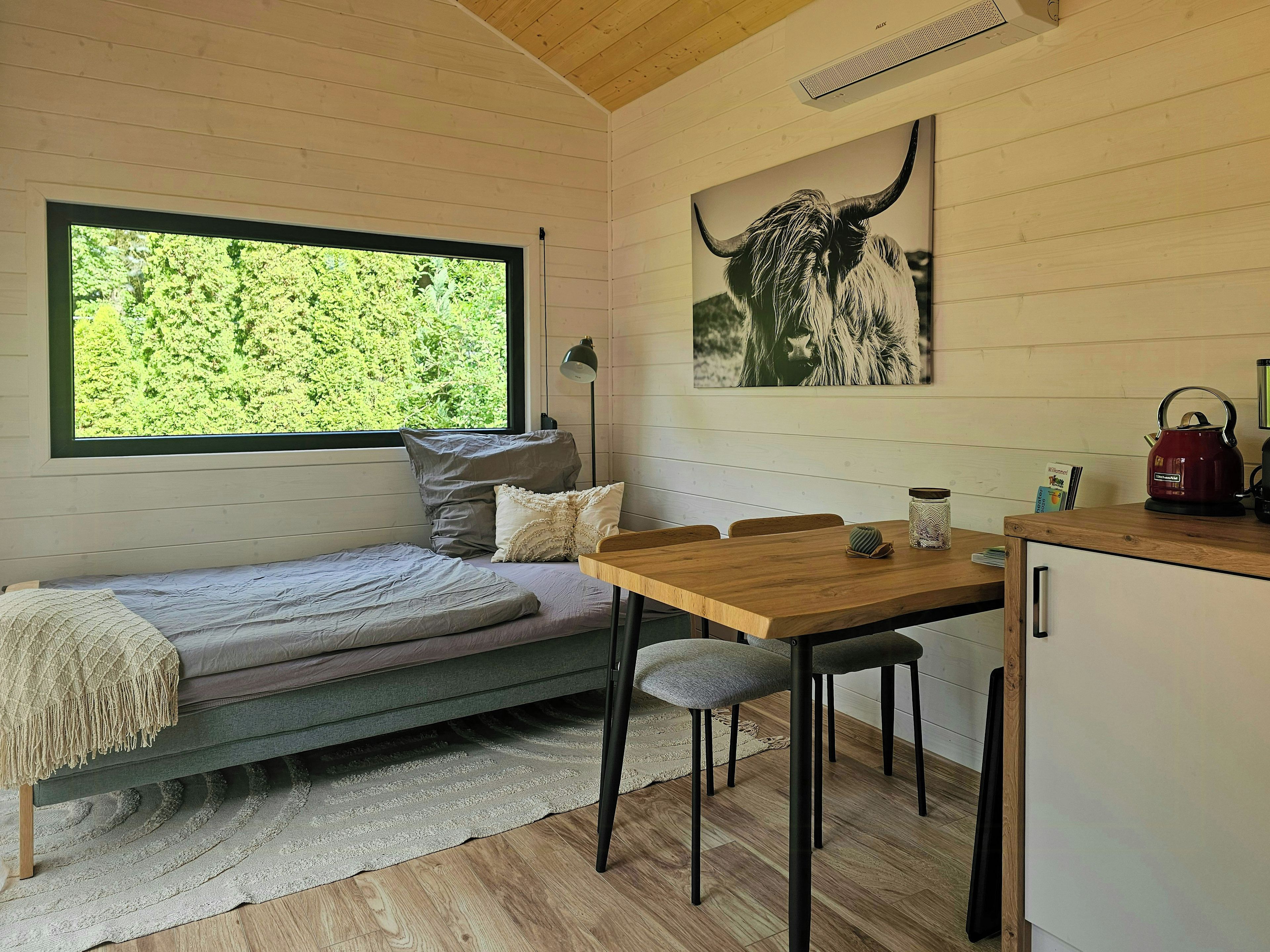 Een knus ingerichte kamer met een bed, een eettafel, stoelen, een waterkoker en een raam met uitzicht op groen.