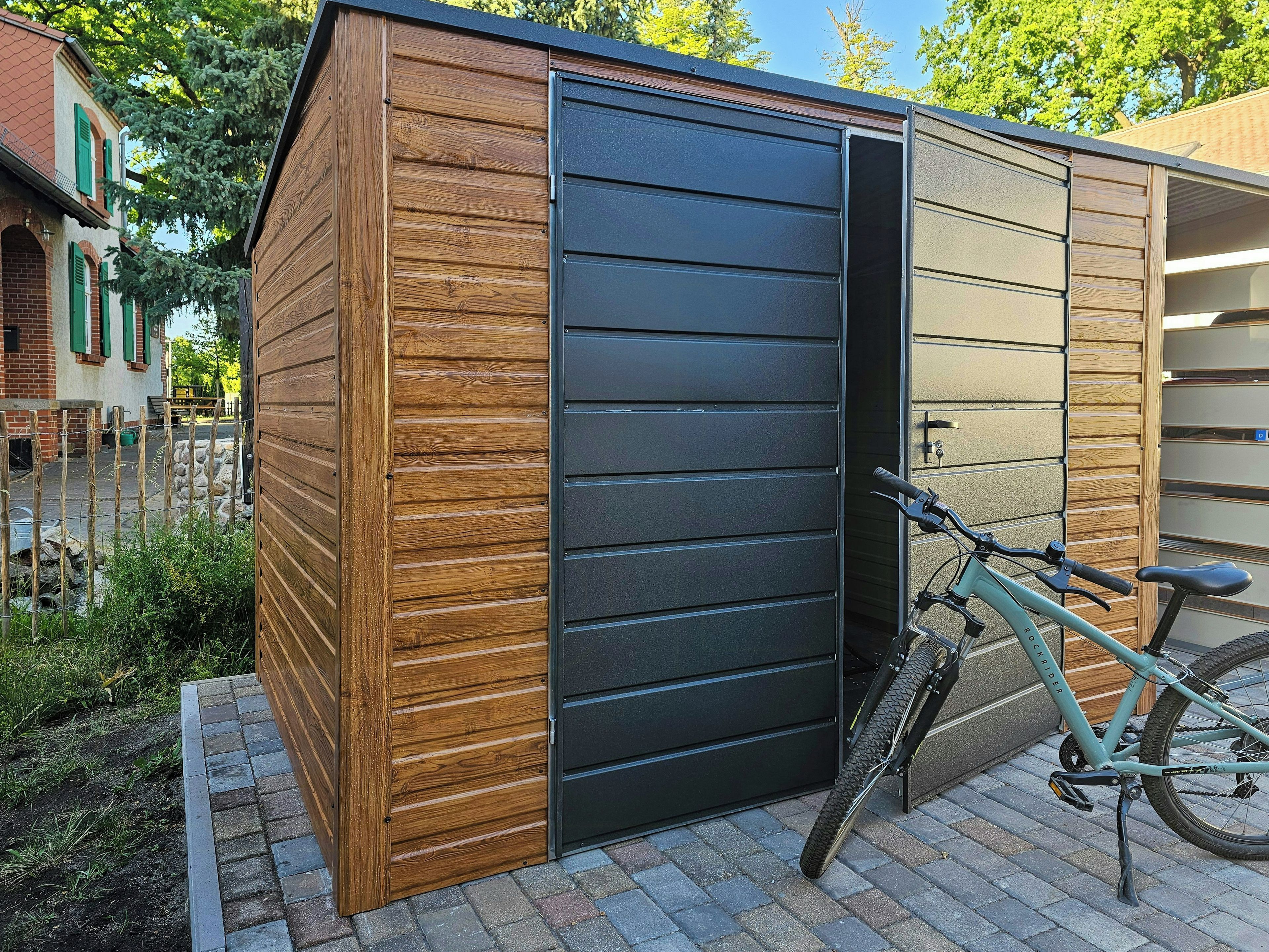 Ein Holzshed mit einer schwarzen Tür, ein Fahrrad davor, eine gepflasterte Fläche mit einem Zaun und Grünpflanzen.