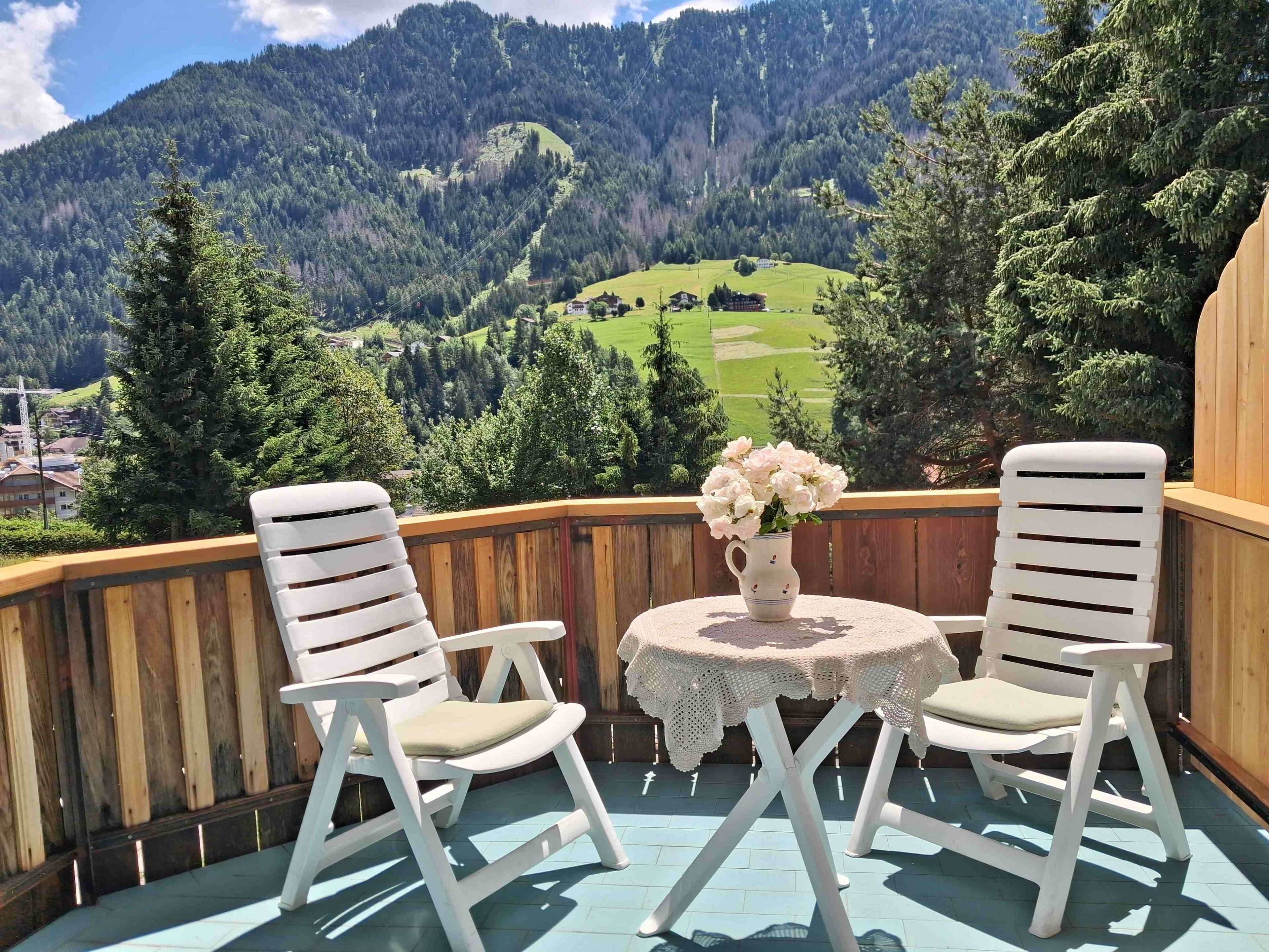 Ein Balkon mit zwei weißen Stühlen und einem Tisch mit Blumen, mit Blick auf eine gebirgige Landschaft.