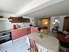 Cocina compacta con estufa, lavamanos y armarios de madera. Hay una zona de comedor con mesa y sillas, y se ve un dormitorio al fondo.