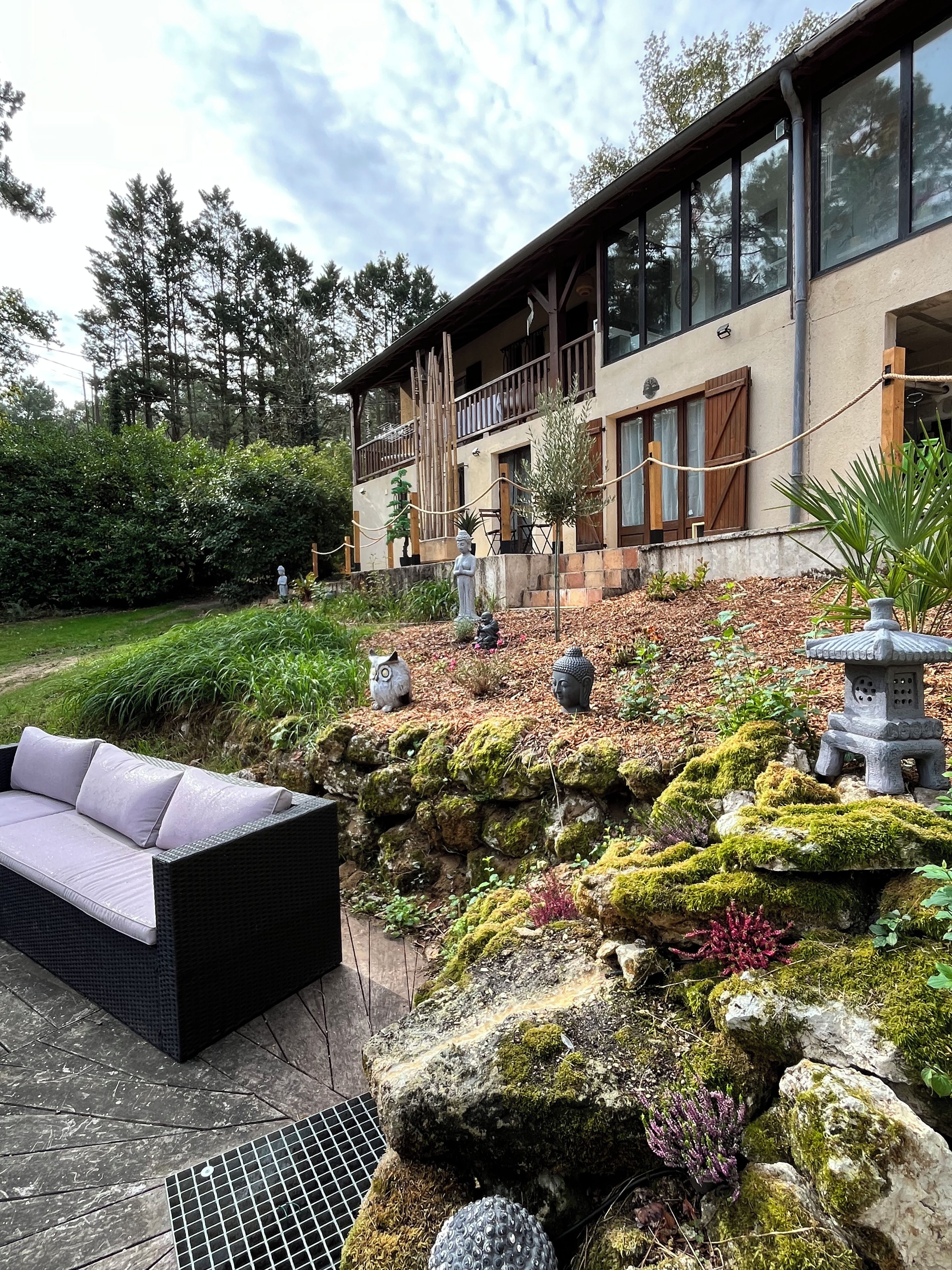 A modern house with a balcony, a stone pathway, and a garden with moss-covered rocks and a Buddha statue.