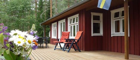 Eine Holzterrasse mit zwei Stühlen, einem Tisch, einer Flagge und einem bewaldeten Hintergrund.