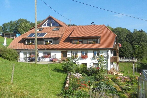 Een huis met twee verdiepingen, een rood pannendak, een tuin met diverse planten en bloemen, en een klein voortuintje.
