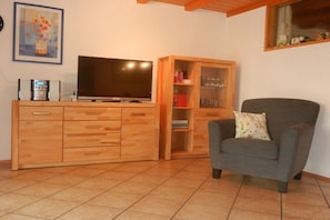 Ein Wohnzimmer mit Fernseher auf einer Holzkommode, einem grauen Sessel und einer Glasvitrine.