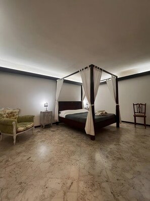 Una cama con dosel y cuatro columnas, una mesita de noche con lámpara, un sillón tapizado de verde, y una silla de madera en un dormitorio con suelo de mármol.