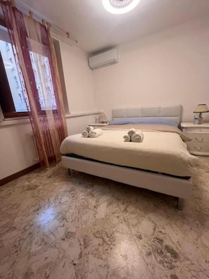 Ein Schlafzimmer mit einem Bett, einem Fenster mit Vorhängen, einer Klimaanlage und einem Nachttisch mit einer Lampe.
