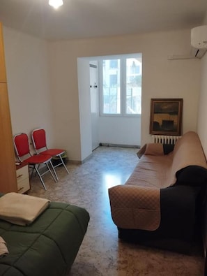 Ein Zimmer mit einem Bett, einem grünen Sofa, einem roten Sessel, einer Heizung und einem Fenster mit Blick auf Gebäude.