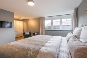 A bedroom with a large bed, a TV mounted on the wall, a window with curtains, and a view of the outside.