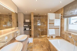 A modern bathroom with a large bathtub, two sinks, a glass shower enclosure, and a towel rack.