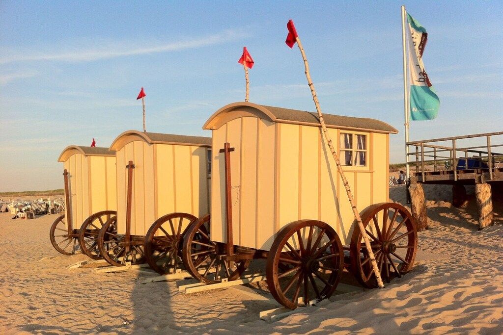 Een strand met twee kleine, witte, gelijkvloerse constructies op bruine wielen, die op wagentjes lijken, en een vlaggenmast met een vlag.