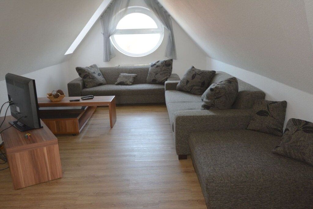 Ein Loft-Wohnzimmer mit großem Fenster, einer Sofagarnitur, einem Holztisch und einem Fernseher auf einem Schrank.