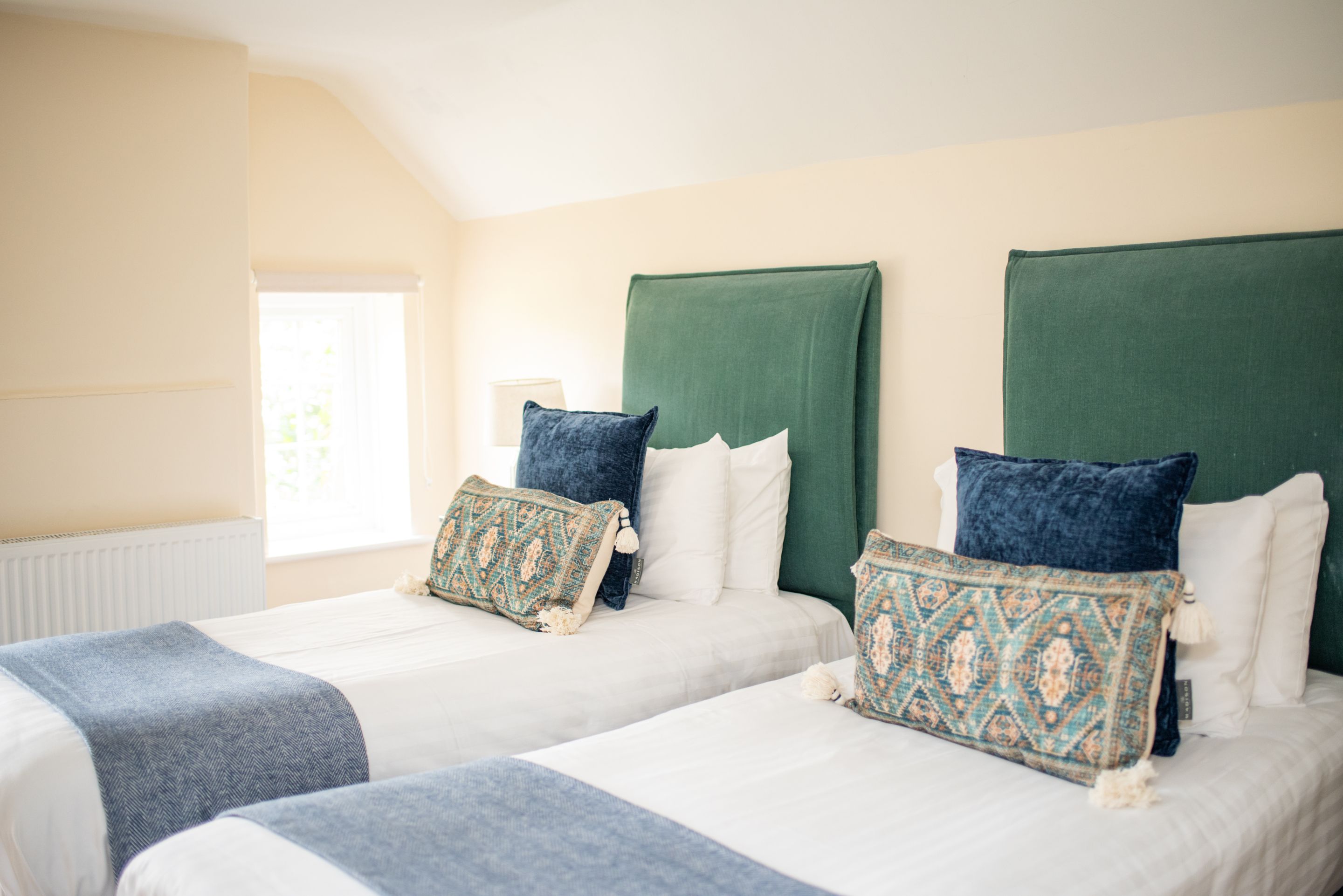Habitación de hotel con dos camas, cada una con cabecero verde, ropa de cama blanca y almohadones estampados.