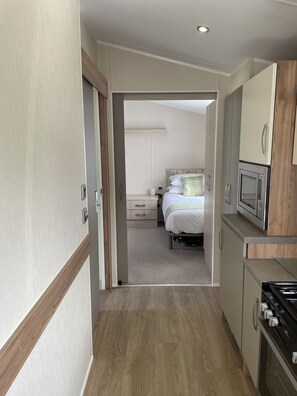 Ein schmaler Flur führt zu einem Schlafzimmer mit Bett, Nachttisch und bei Nachtlicht. Eine Küchenzeile mit Herd und Mikrowelle ist rechts sichtbar.