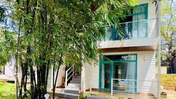 A two-story building with a balcony, surrounded by lush greenery and a wet ground, suggesting recent rain.