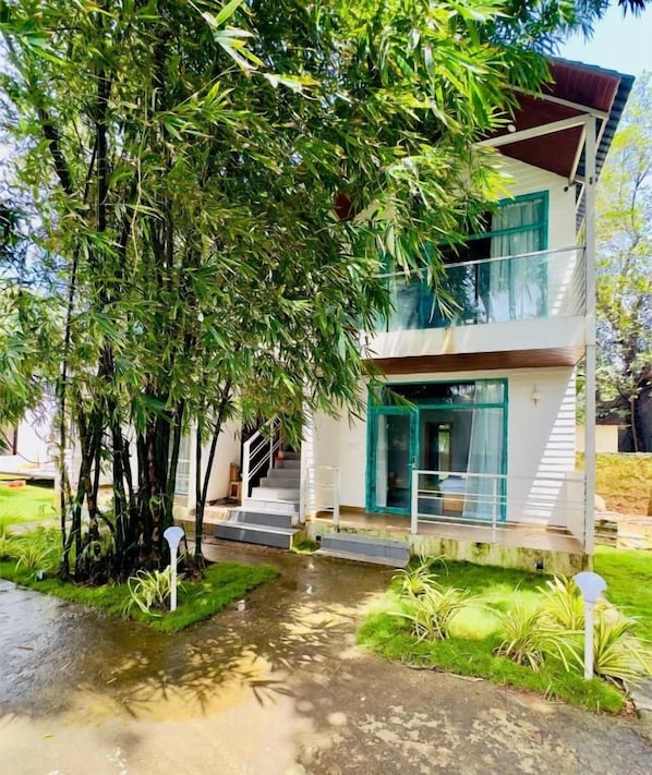 Un edificio de dos plantas con balcón, rodeado de abundante vegetación y suelo húmedo, lo que sugiere lluvia reciente.