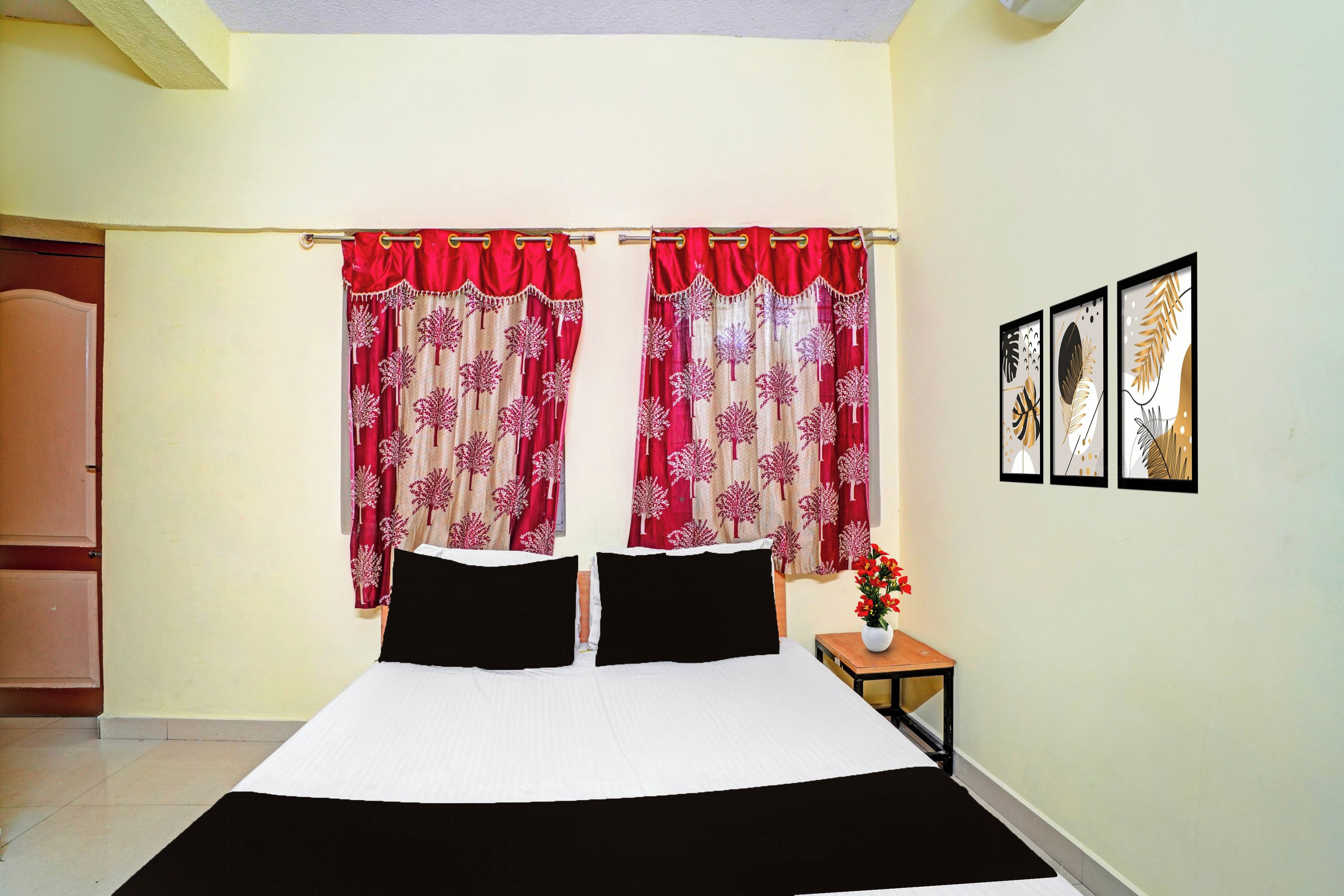 Une chambre à coucher avec un lit, deux oreillers, une table de chevet ornée d’un vase de fleurs et une porte située à gauche.