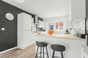 A modern kitchen with white cabinets, a central island with stools, a microwave, and a fruit bowl. - The Harmony House at Honor Estate (Telford)