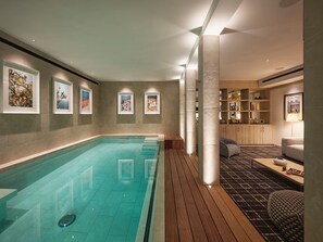 Ein moderner, überdachter Schwimmbereich mit Holzboden, einer Sitzecke mit Sofa und Sesseln sowie gerahmten Kunstwerken an den Wänden.