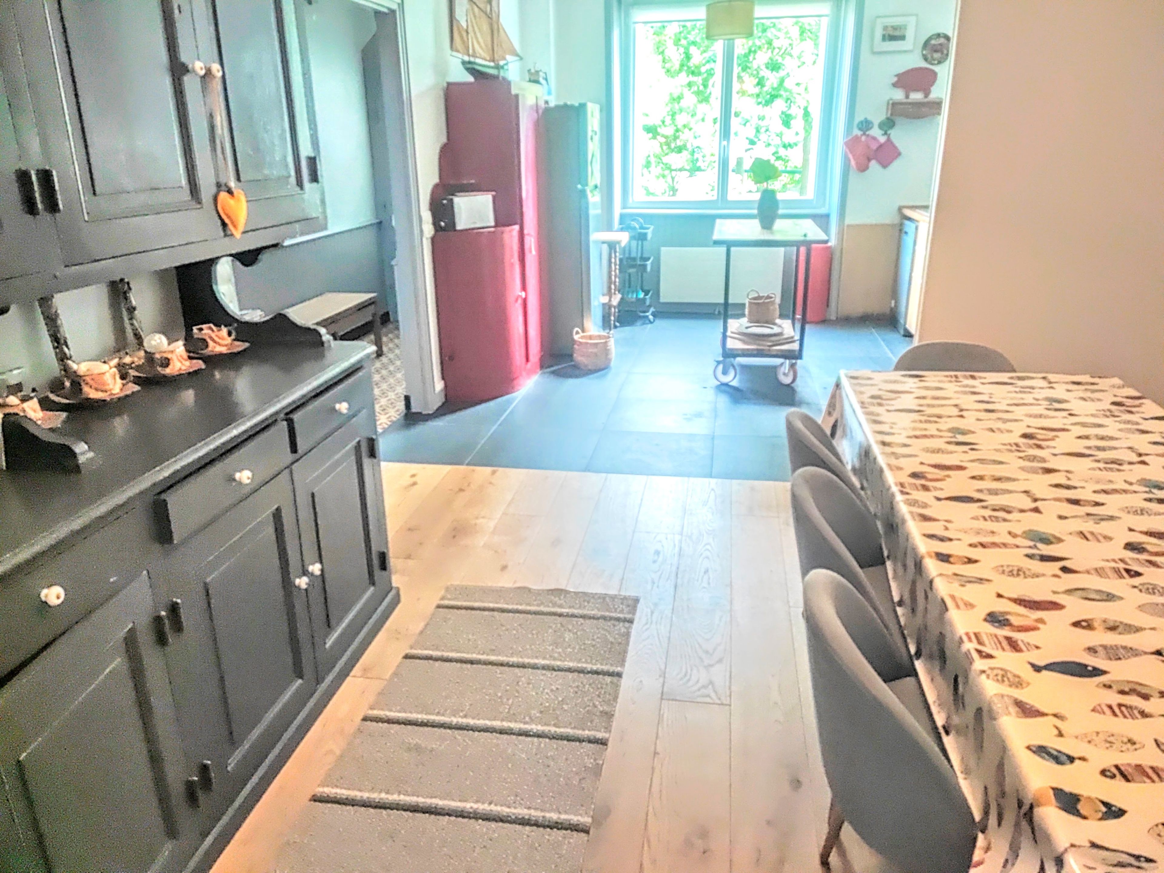A kitchen with a dining area, a window with a view of greenery, and a table with a fish pattern tablecloth.