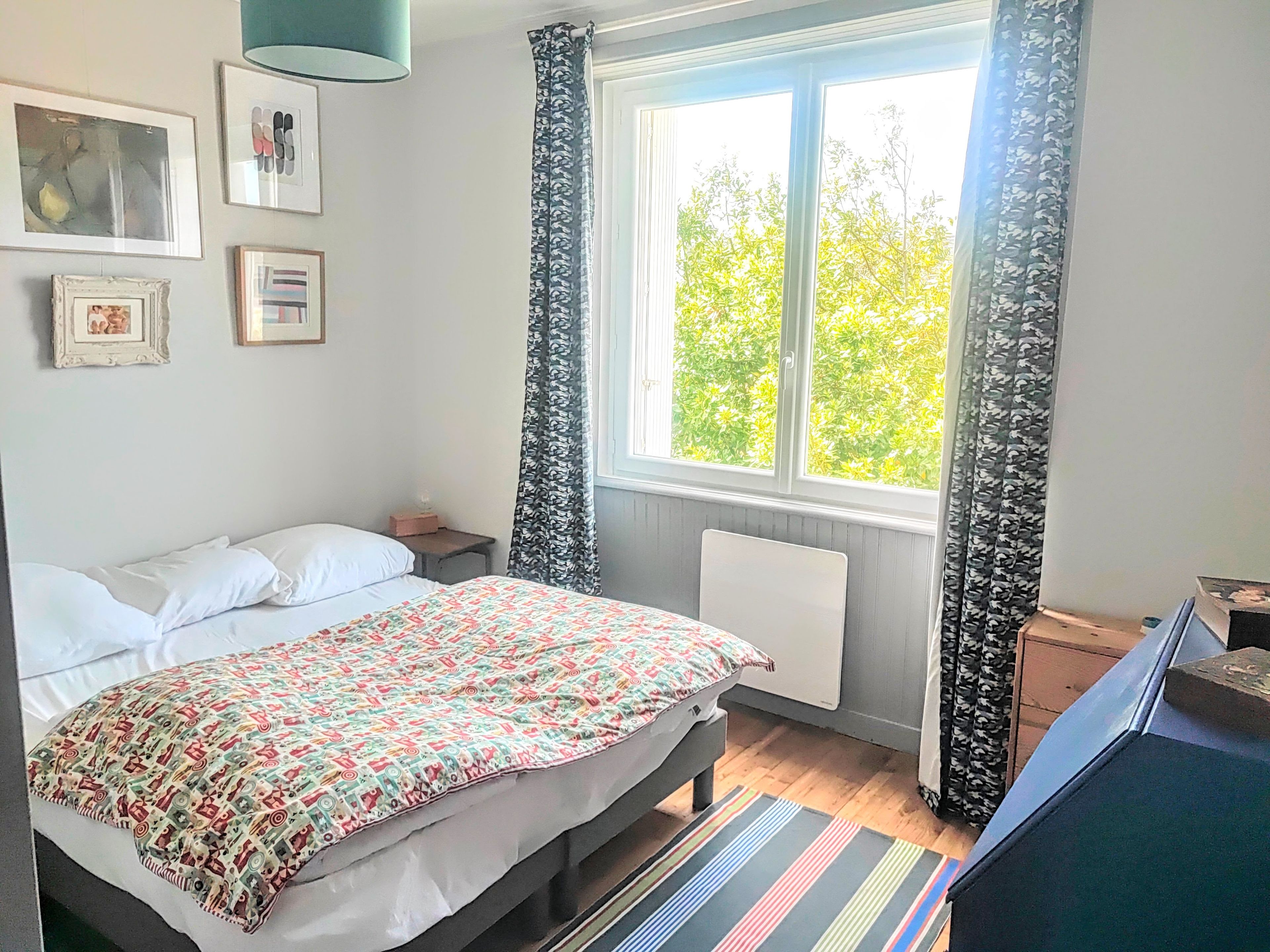 Un dormitorio con una cama, una mesita de noche, un radiador, una ventana con cortinas y una obra de arte en la pared.