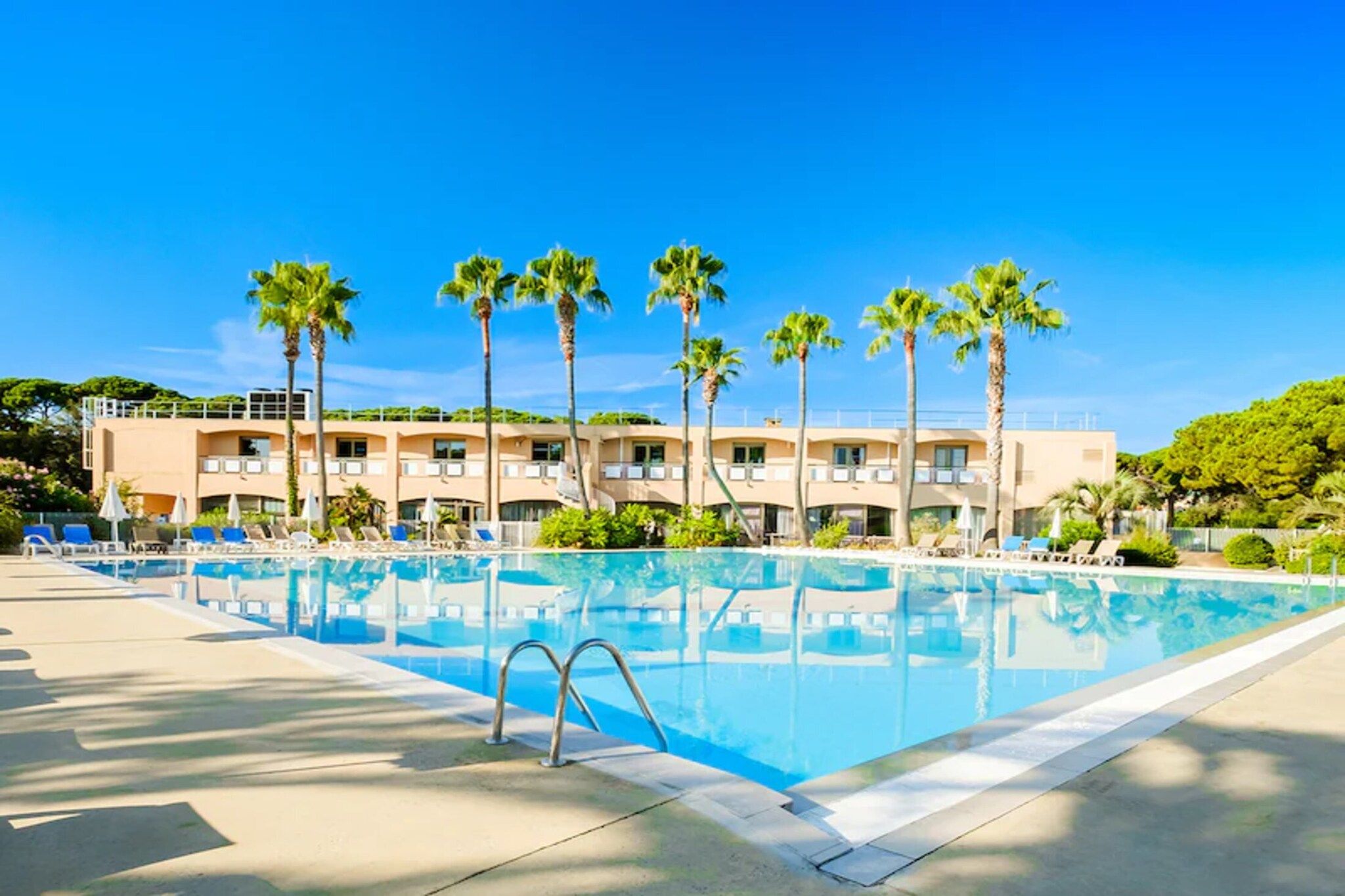 A swimming pool with lounge chairs and palm trees, surrounded by a building with multiple balconies.