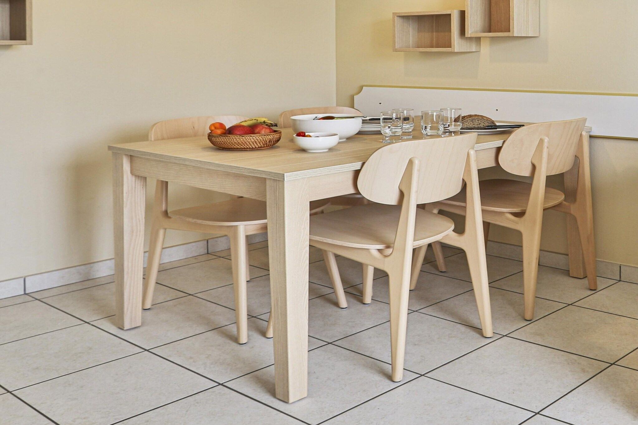 Une table à manger avec quatre chaises, un panier de fruits et un saladier blanc.