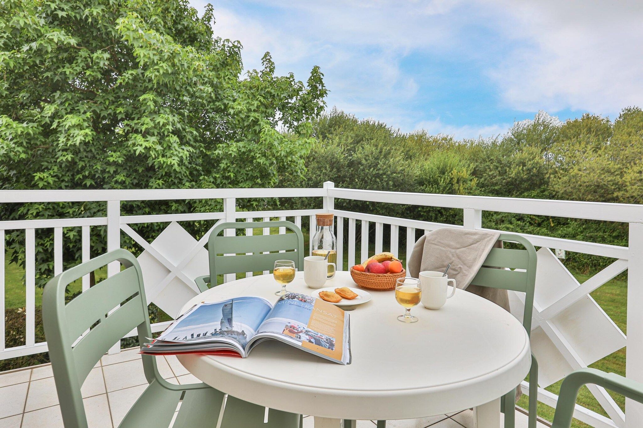 Une table ronde avec un magazine, un verre de vin, une assiette de fruits et une tasse sur un balcon doté de balustrades blanches et de chaises vertes.