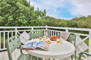 Une table ronde avec un magazine, un verre de vin, une assiette de fruits et une tasse sur un balcon doté de balustrades blanches et de chaises vertes.