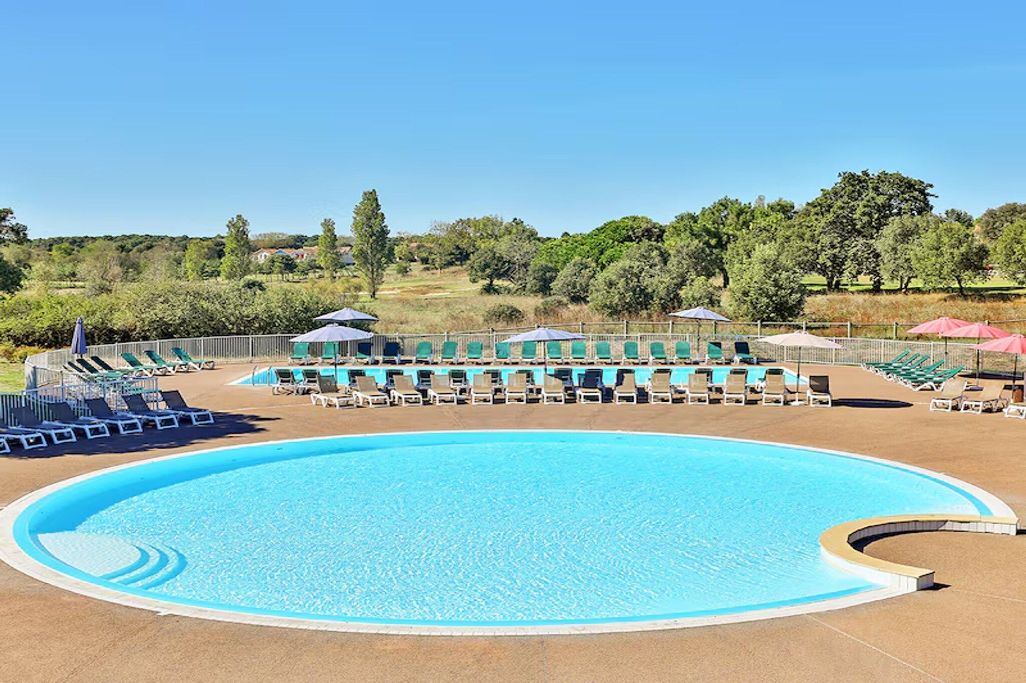 Une grande piscine extérieure, dotée de nombreux chaises longues et parasols, et entourée d’une clôture et d’arbres.