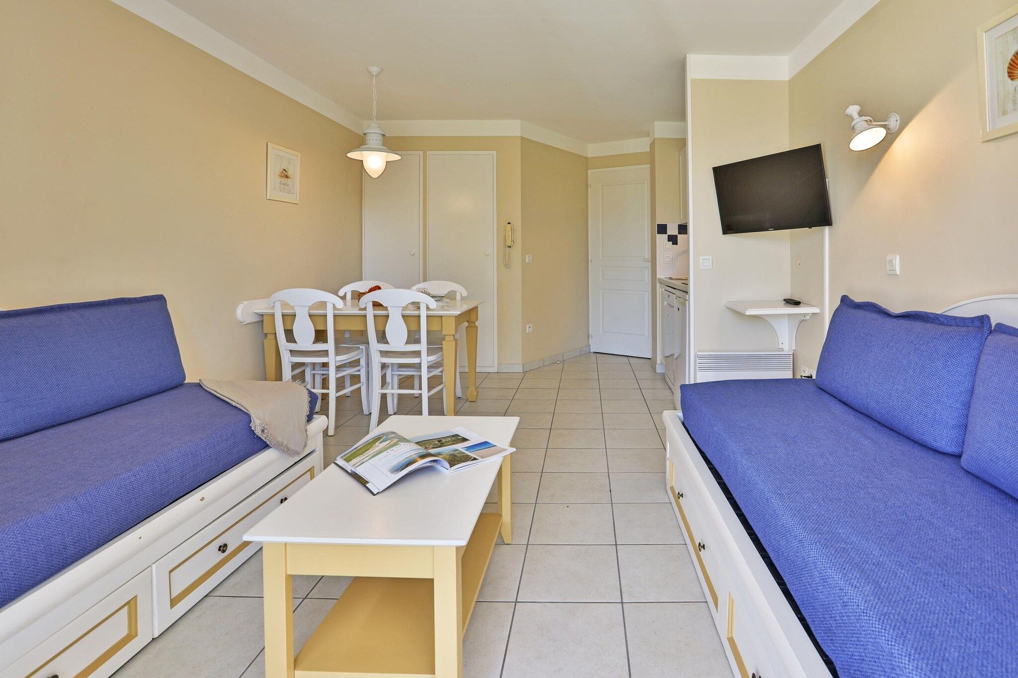 Un espace de vie compact comprenant un canapé, une table à manger avec des chaises, un téléviseur fixé au mur et un coin cuisine à l’arrière-plan.