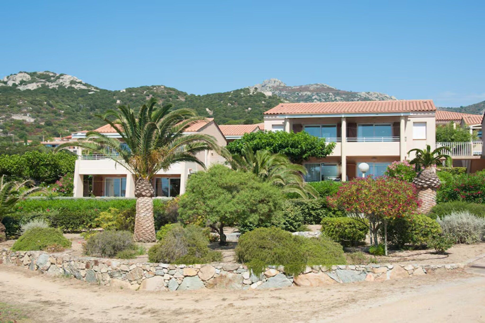 A two-story building with a red-tiled roof, surrounded by landscaped gardens with various shrubs and a palm tree.