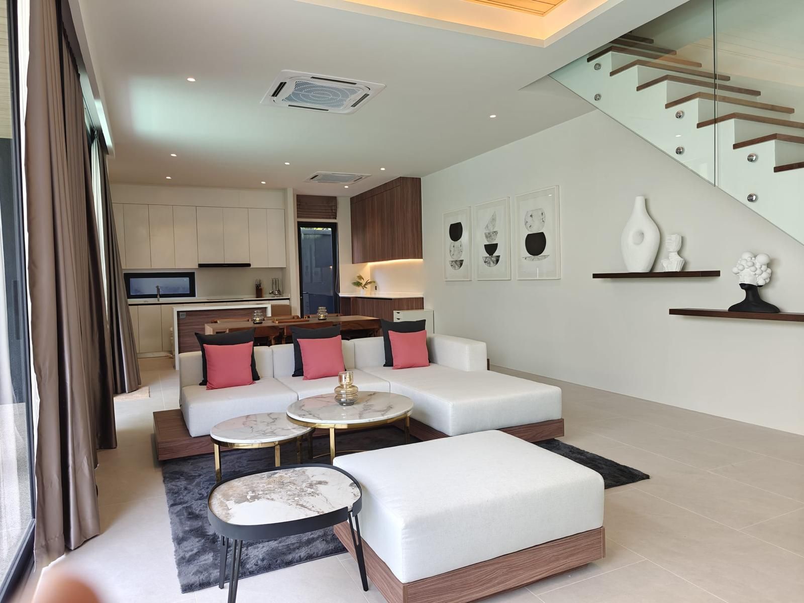 Ein modernes Wohnzimmer mit Sofa, Hocker und Couchtisch, im Hintergrund eine Küche und eine Treppe.