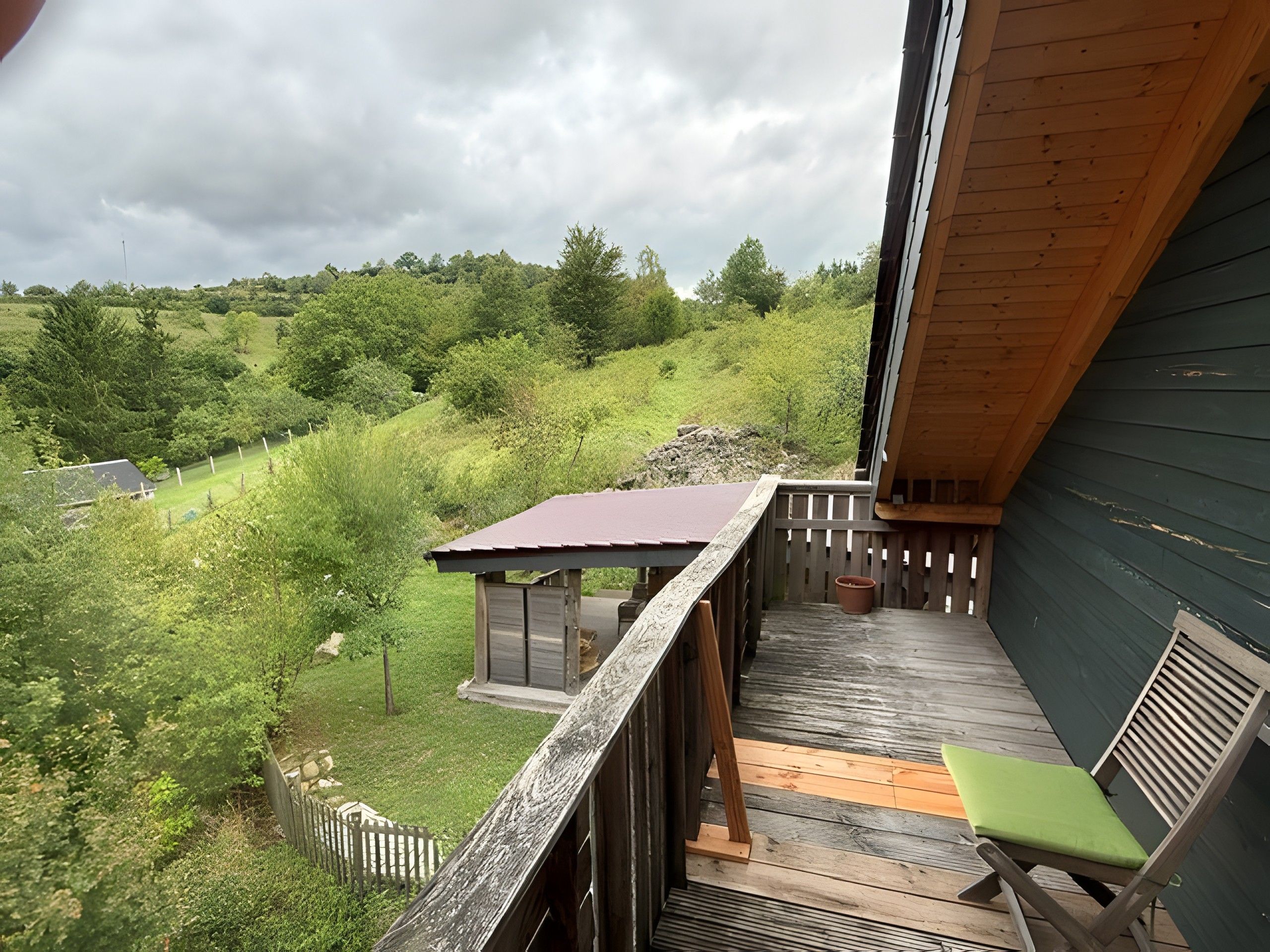 Una terraza de madera con vistas a un paisaje verde, una pequeña estructura con techo rojo y una silla verde.