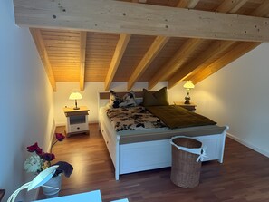A bedroom with a sloped ceiling, a bed with a floral bedspread, two bedside tables with lamps, and a wicker basket. - Holiday Home 'Ferienwohnung Lou' with Shared Garden, Balcony and Wi-Fi (Beverungen)
