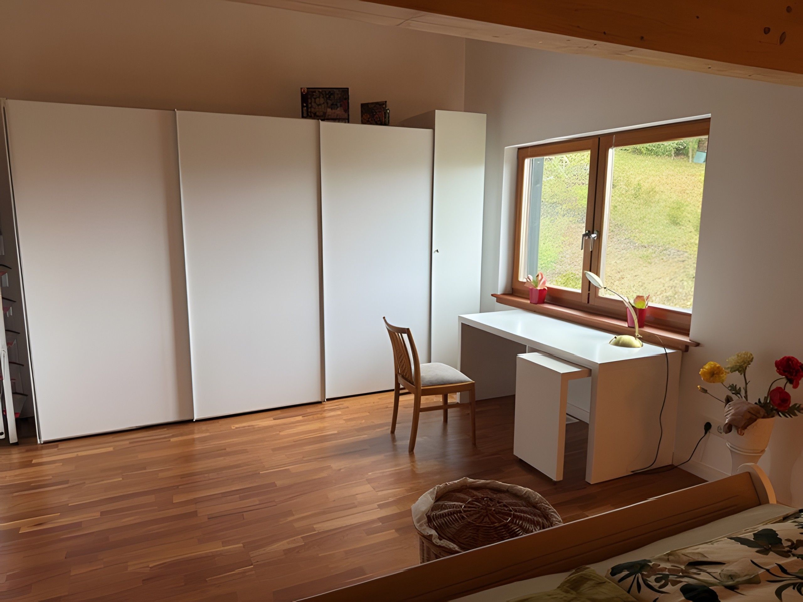 A room with a wooden floor, a desk with a chair, a window with a view of greenery, and a bed with a floral bedspread.