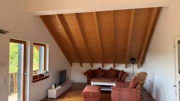 Habitación estilo loft con techo inclinado, piso de madera, sofá, sillón y televisor.