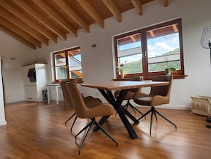 A dining area with a wooden table and chairs, a kitchen in the background, and large windows with a view of greenery. - Holiday Home 'Ferienwohnung Lou' with Shared Garden, Balcony and Wi-Fi (Beverungen)