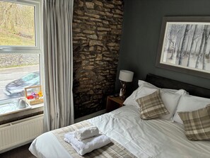 Une chambre d’hôtel avec un mur en pierre, un lit recouvert de draps blancs, une fenêtre avec des rideaux, une table de chevet avec une lampe et un tableau encadré accroché au mur.