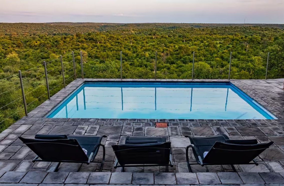 Una piscina rectangular con superficie de agua azul cristalina, rodeada de baldosas de piedra. Hay tres tumbonas dispuestas frente a la piscina, y una cerca de metal delimita el espacio. Al fondo, un paisaje verde y exuberante.