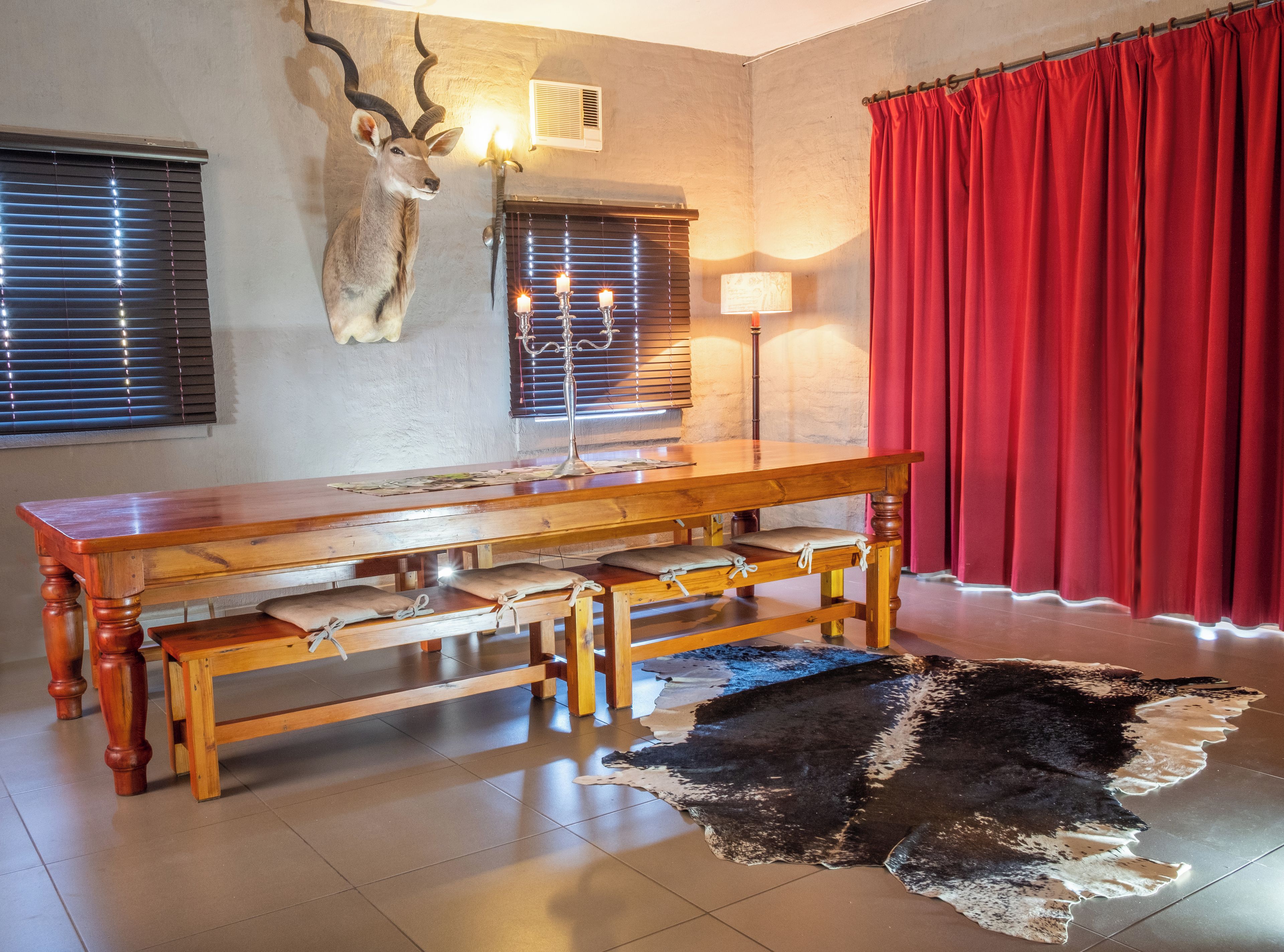 A room with a wooden table, benches, a fur rug, a wall-mounted animal head, and a red curtain.