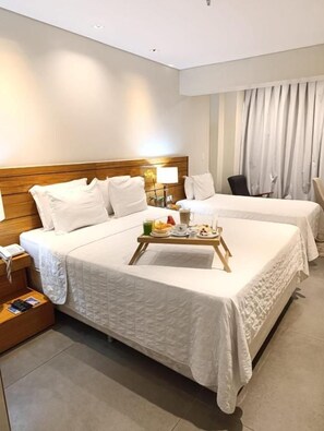 Habitación de hotel con dos camas, cabecero de madera, mesitas de noche con lámparas y una bandeja con alimentos para el desayuno.