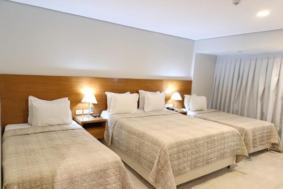 Habitación de hotel con dos camas, cabecero de madera, lámparas de noche y una ventana con cortinas.
